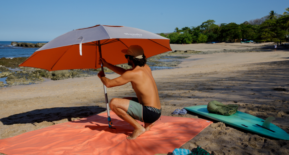 Dad seats a deep-set beach anchor with a hammer-in motion as a UPF 55+ silver canopy opens; kids play on a breezy shoreline.