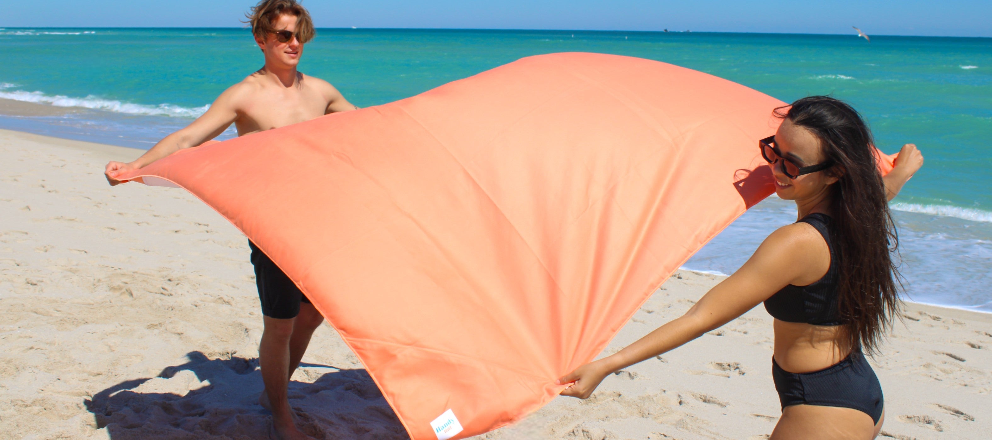 A man and a woman are setting up the Handy Beach Blanket on the beach sand