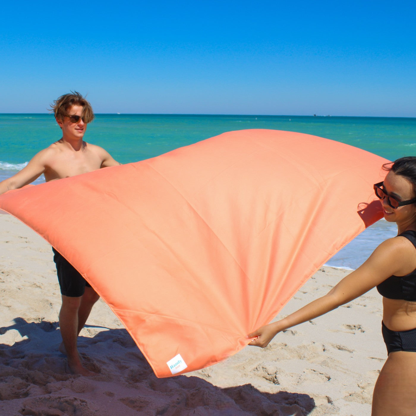 A man and a woman are setting up the Handy Beach Blanket on the beach sand