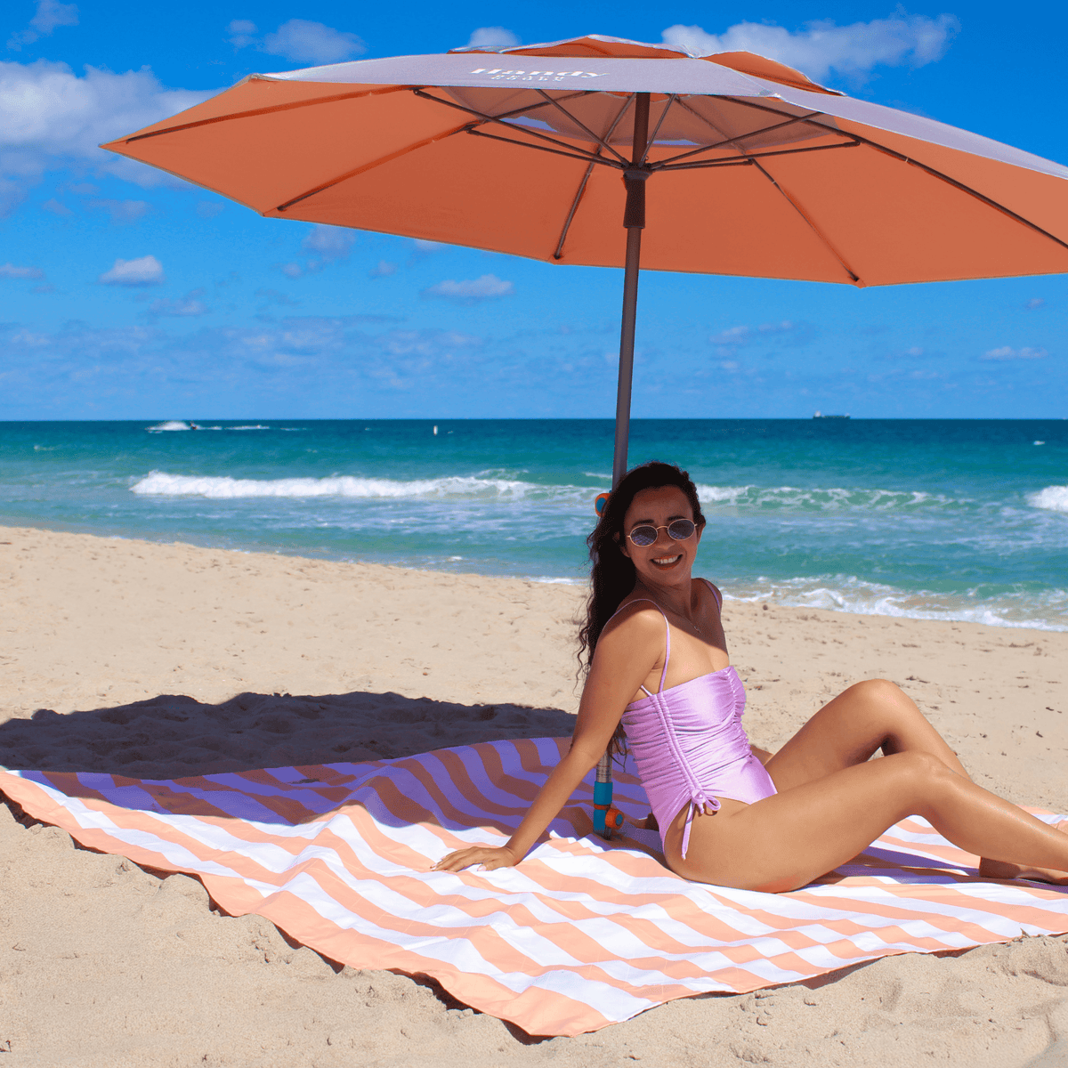 Woman sitting under a silver & orange umbrella on a beach with ocean in the background