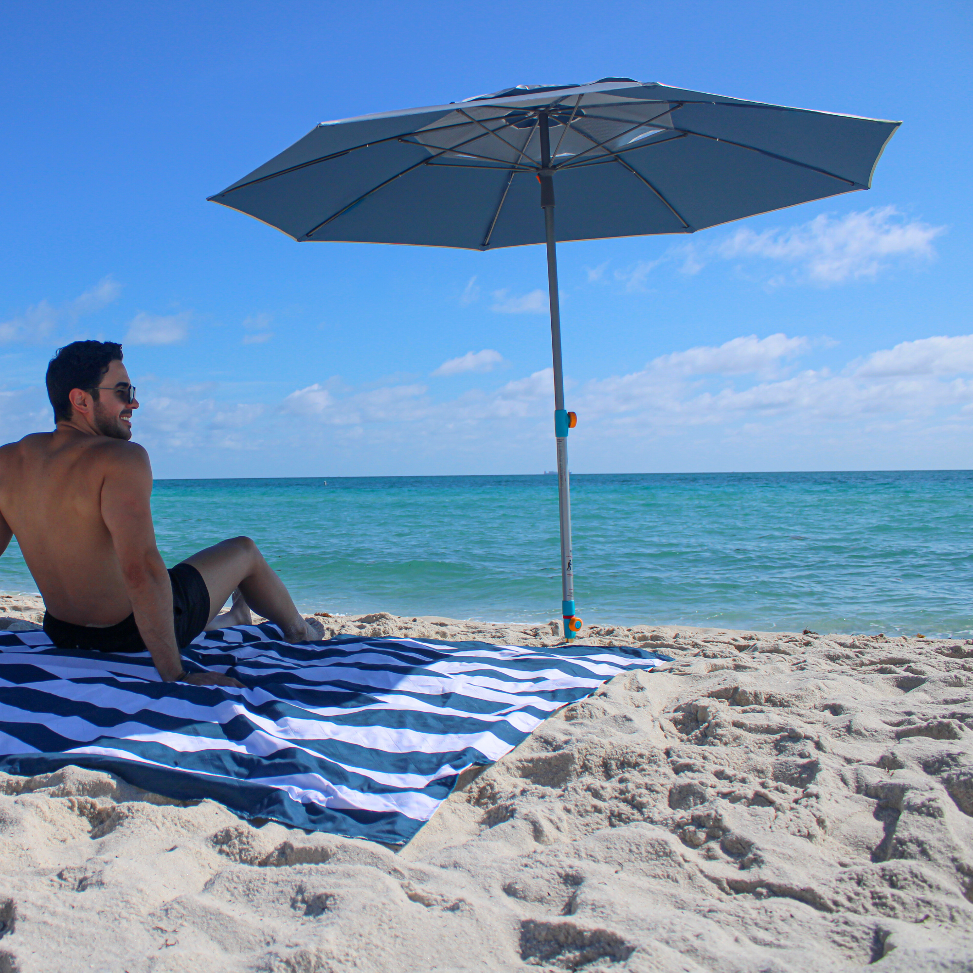 Silver and blue Handy Beach Umbrella providing deep shade for a couple relaxing on the beach near the ocean.