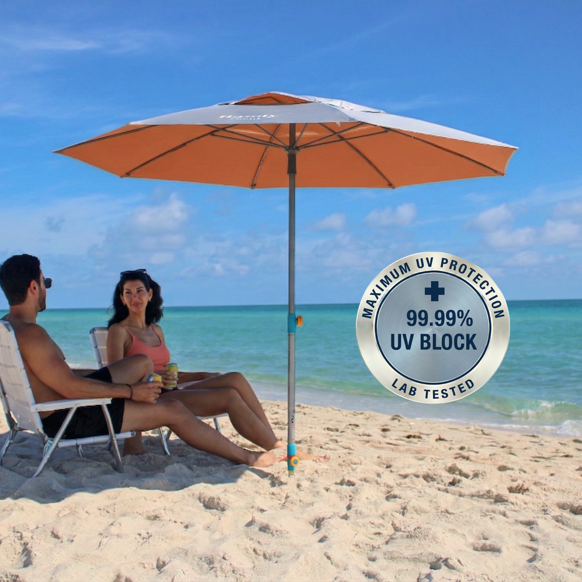 A silver and orange beach umbrella stands on a sandy beach with two people sitting underneath in white lounge chairs. The turquoise ocean and blue sky are in the background, and a circular badge on the right reads 'MAXIMUM UV PROTECTION 99.99 % UV BLOCK"