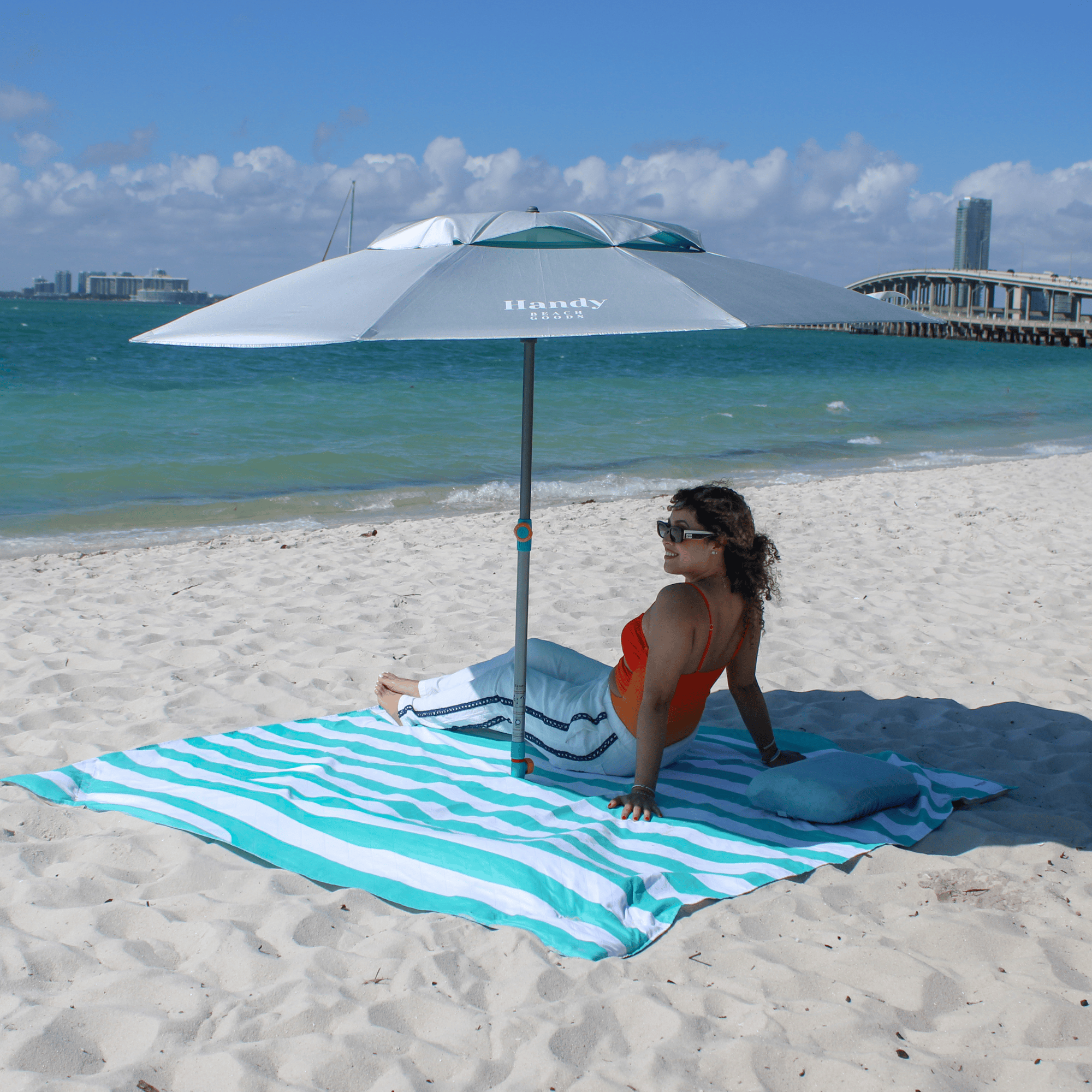 Woman sitting under a large umbrella on a beach blanket in teal stripes with ocean and pier in the background