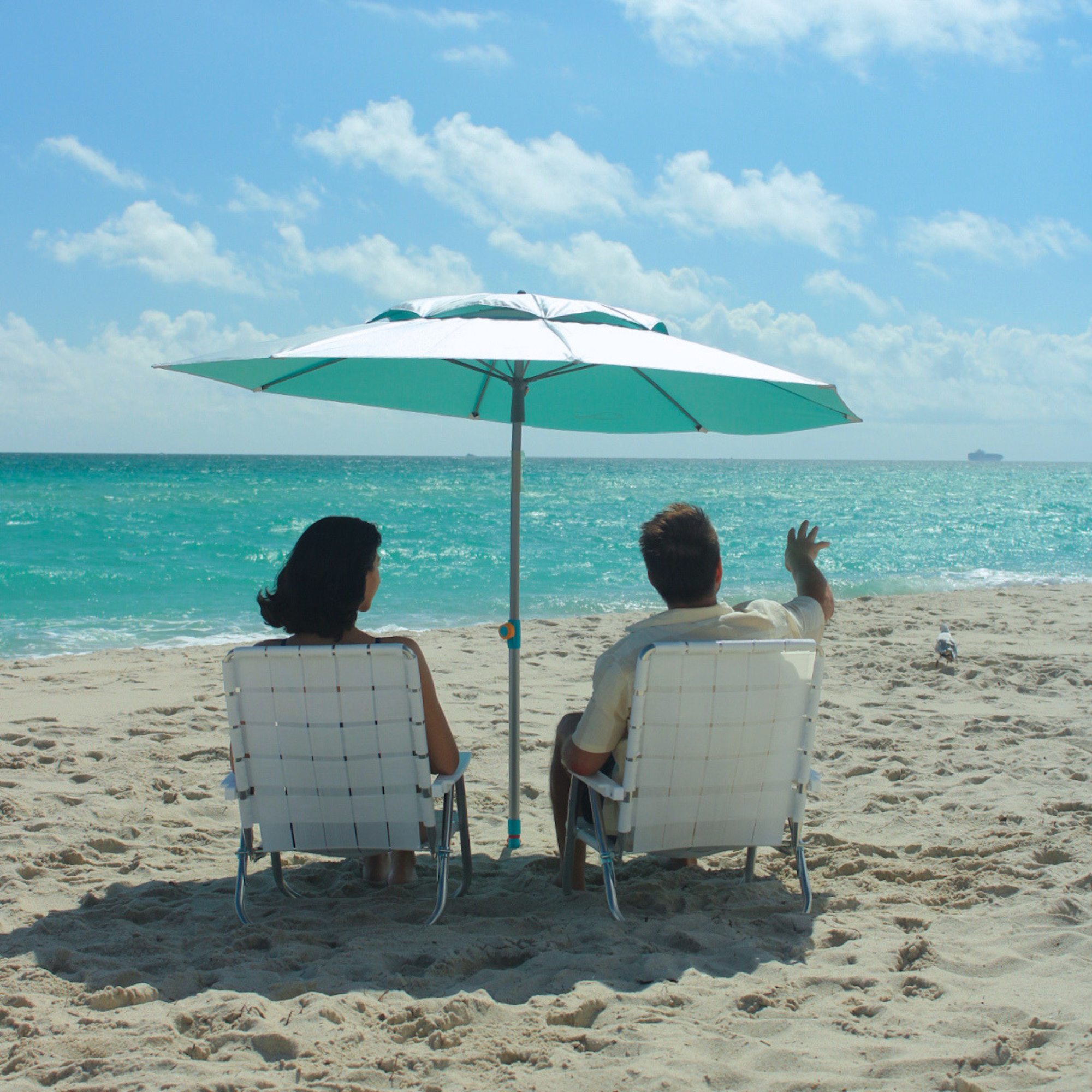 Two people sitting under a top-rated  silver and teal umbrella on a sandy beach with ocean view