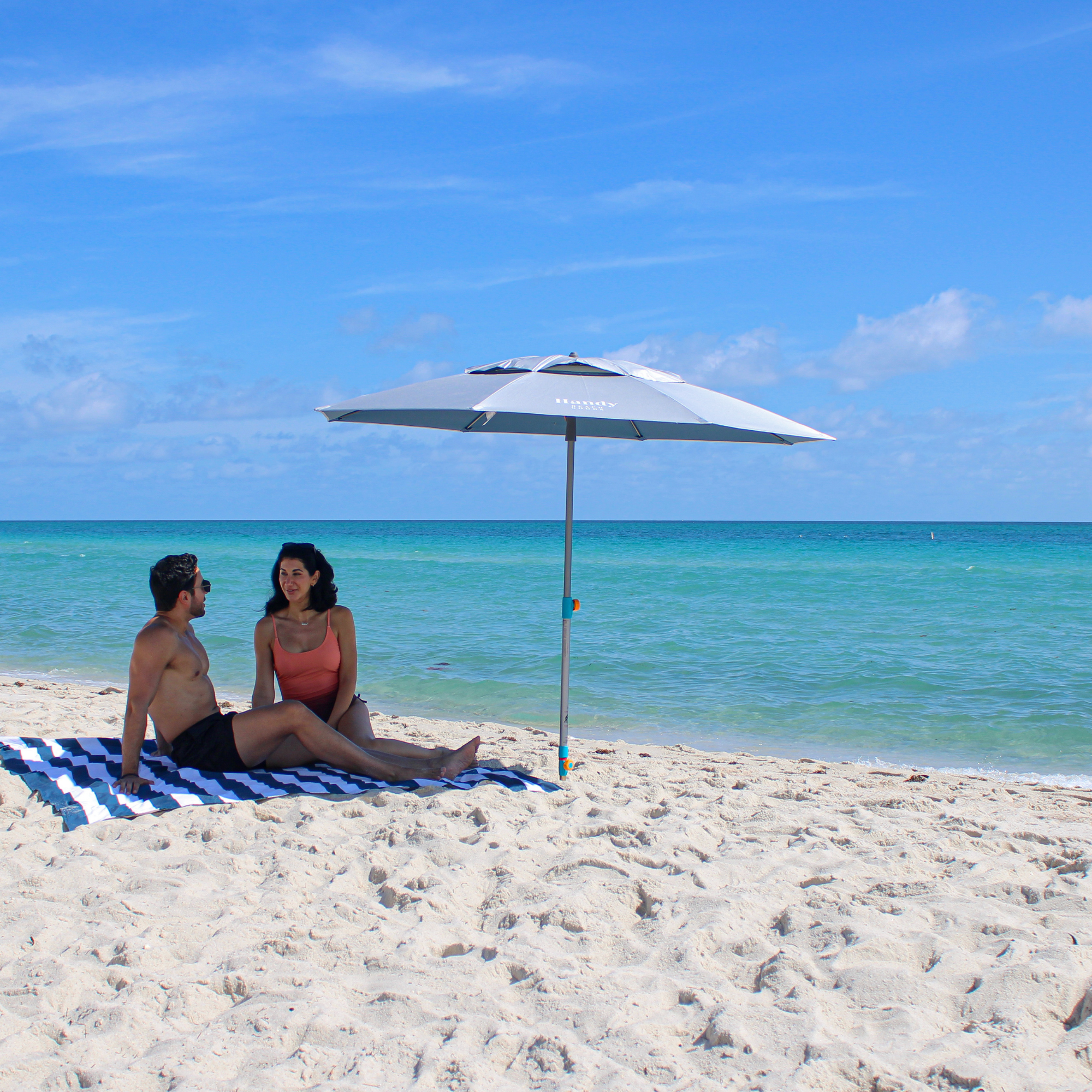 Silver and blue Handy Beach Umbrella providing deep shade for a couple relaxing on the beach near the ocean.