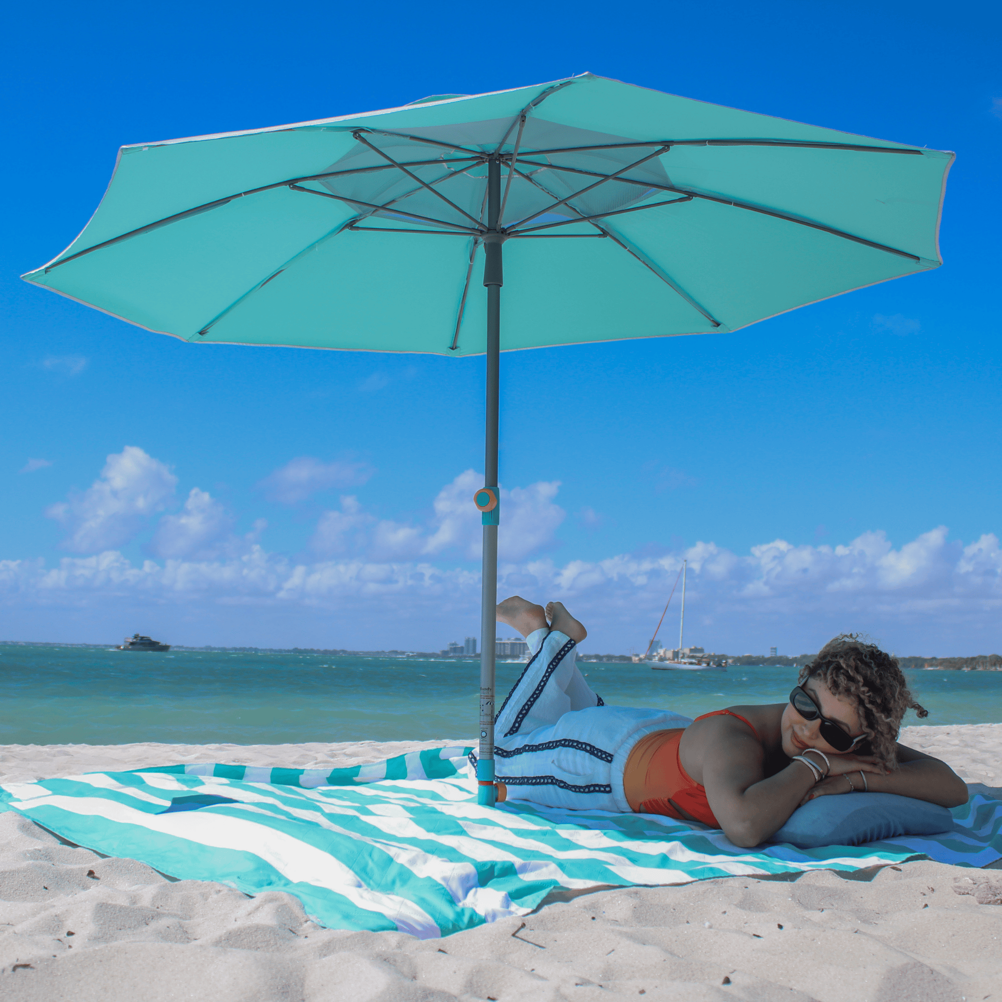 Silver and teal Handy Beach Umbrella providing deep shade for a woman relaxing on the beach near the ocean. teal umbrella system