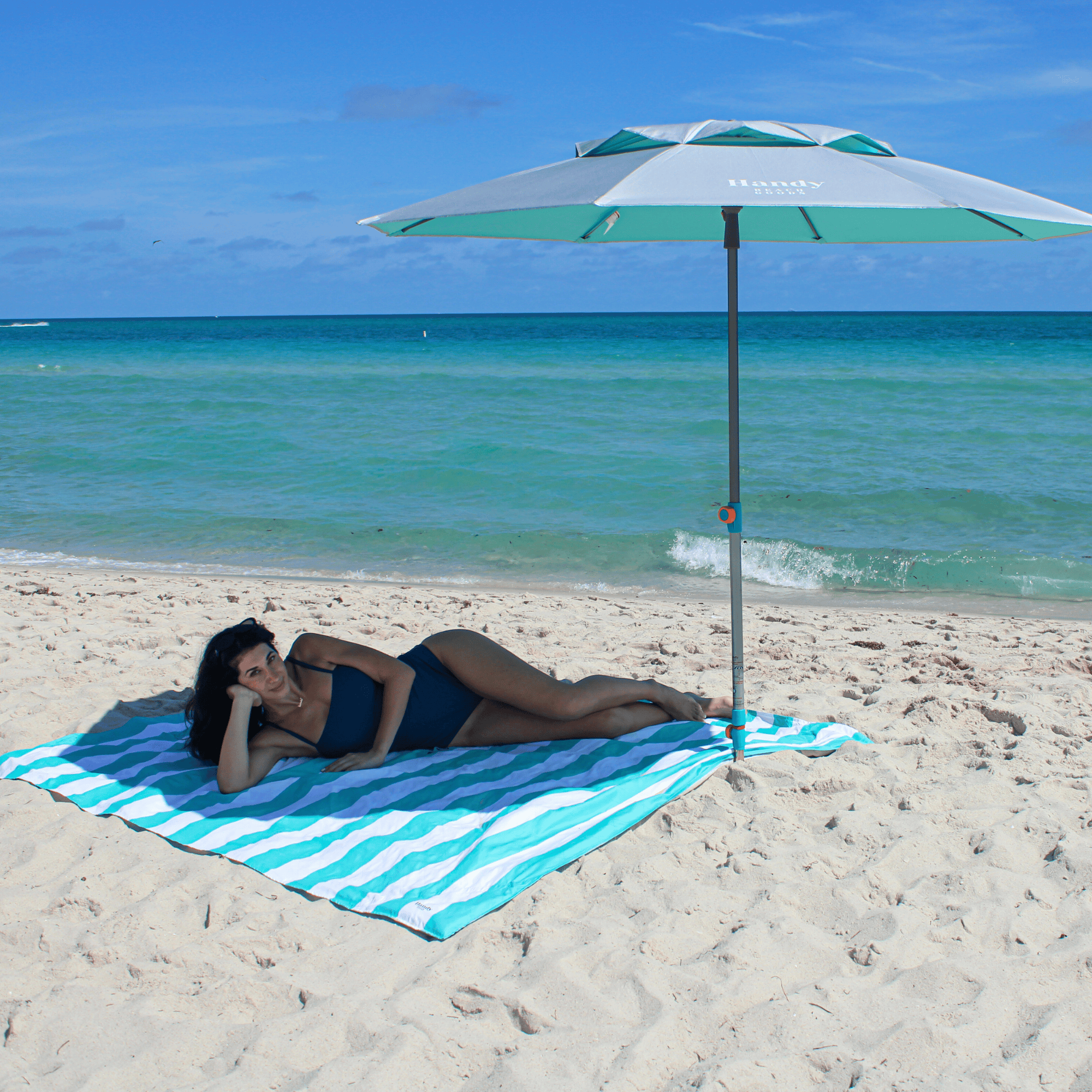 Silver and teal Handy Beach Umbrella providing deep shade for a millennial woman relaxing on the beach near the ocean. teal umbrella system